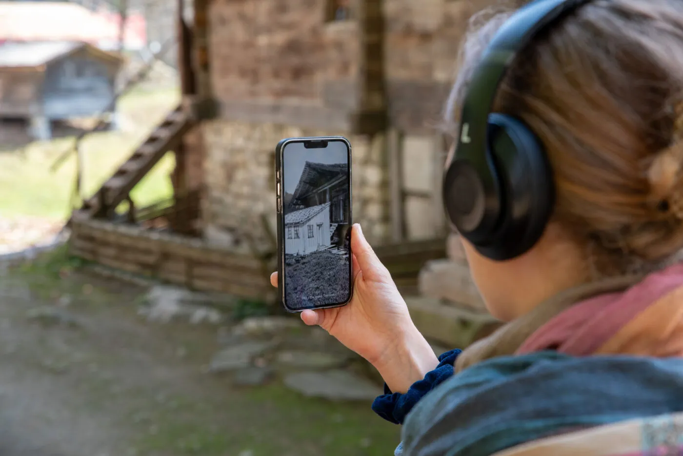 Eine Frau mit Kopfhörer blickt auf ein Smartphone, das in der AR-Anwendung den alten Restaurantanbau des Hauses zeigt